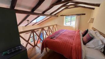 Ein Schlafzimmer mit Holzboden, einem Bett mit Kissen, einem Fenster mit Vorhängen und Blick ins Freie.