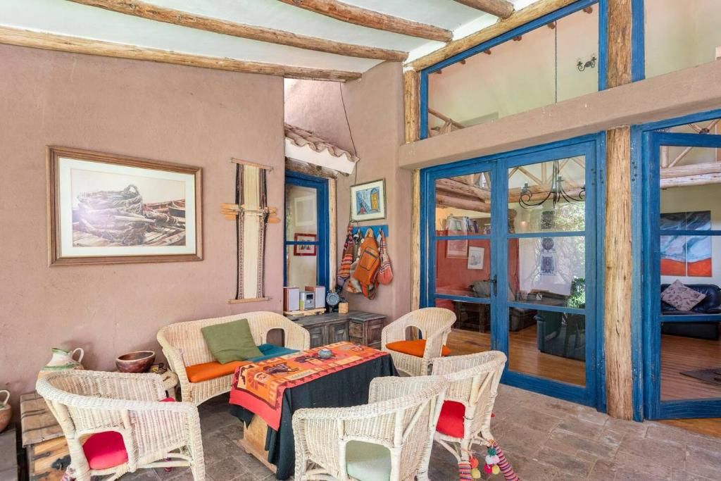 Une pièce meublée de meubles en osier, une table recouverte d’une nappe colorée et une grande fenêtre ornée de bordures bleues.