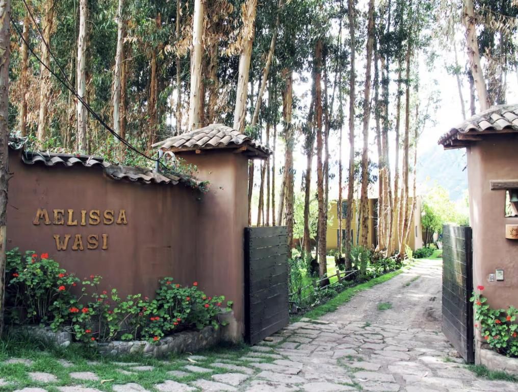 A gated entrance with a nameplate reading 'AELISSA WASI' and a cobblestone path leading through a garden with red flowers.
