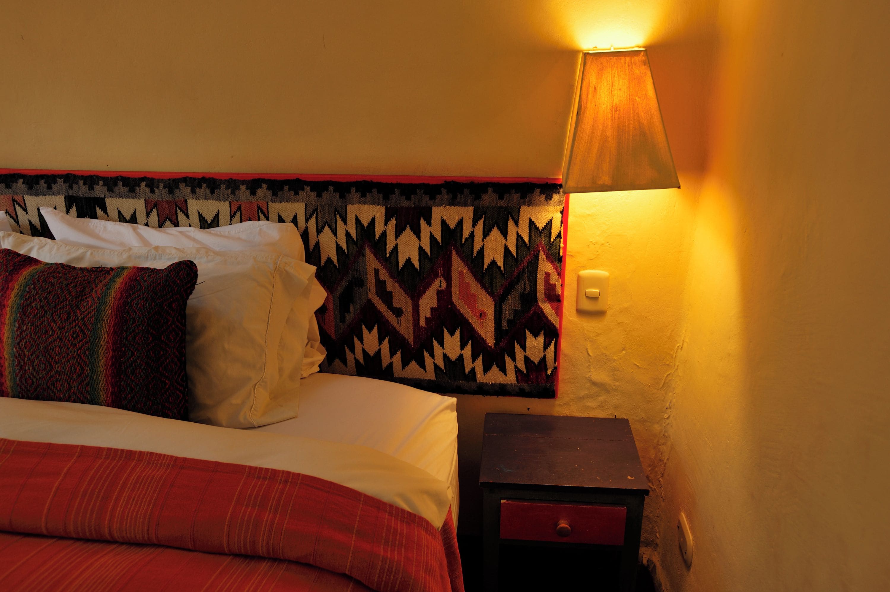 Cama com roupa de cama branca e vermelha, cabeceira estampada e mesa de cabeceira com abajur.