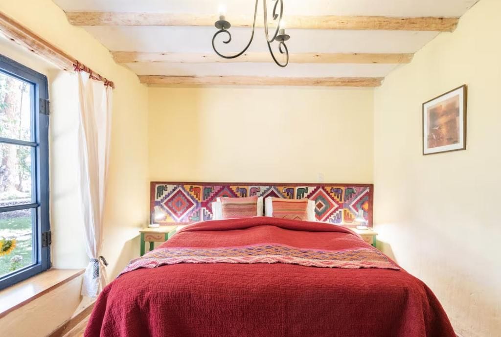A room with a bed, a window with curtains, a patterned headboard, and a chandelier.