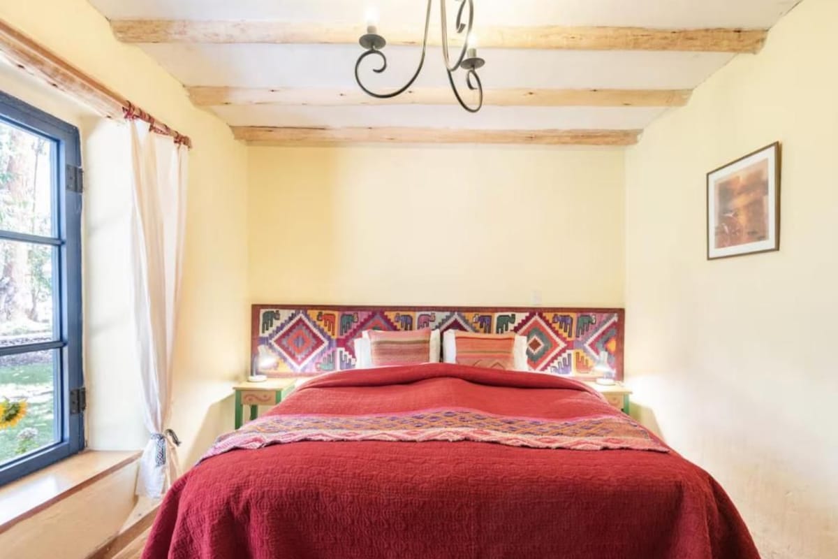 Una habitación con cama, ventana con cortinas, cabecero estampado y candelabro.