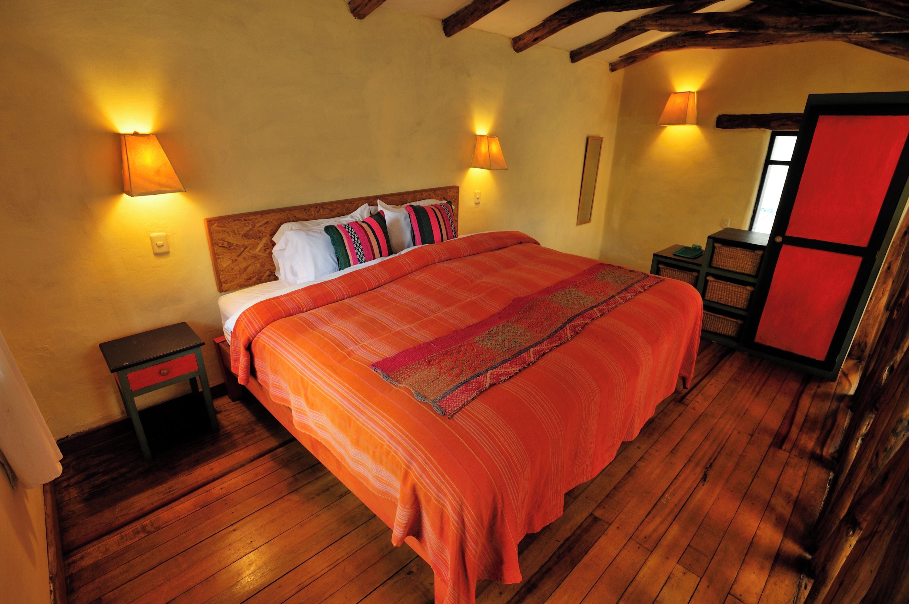 Una habitación con piso de madera, una cama con colcha naranja, dos mesitas de noche y una puerta roja.