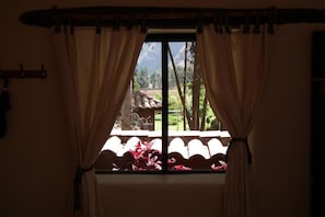 A window with curtains, a view of a garden, and a mountain in the background. - Melissa Wasi (Písac)