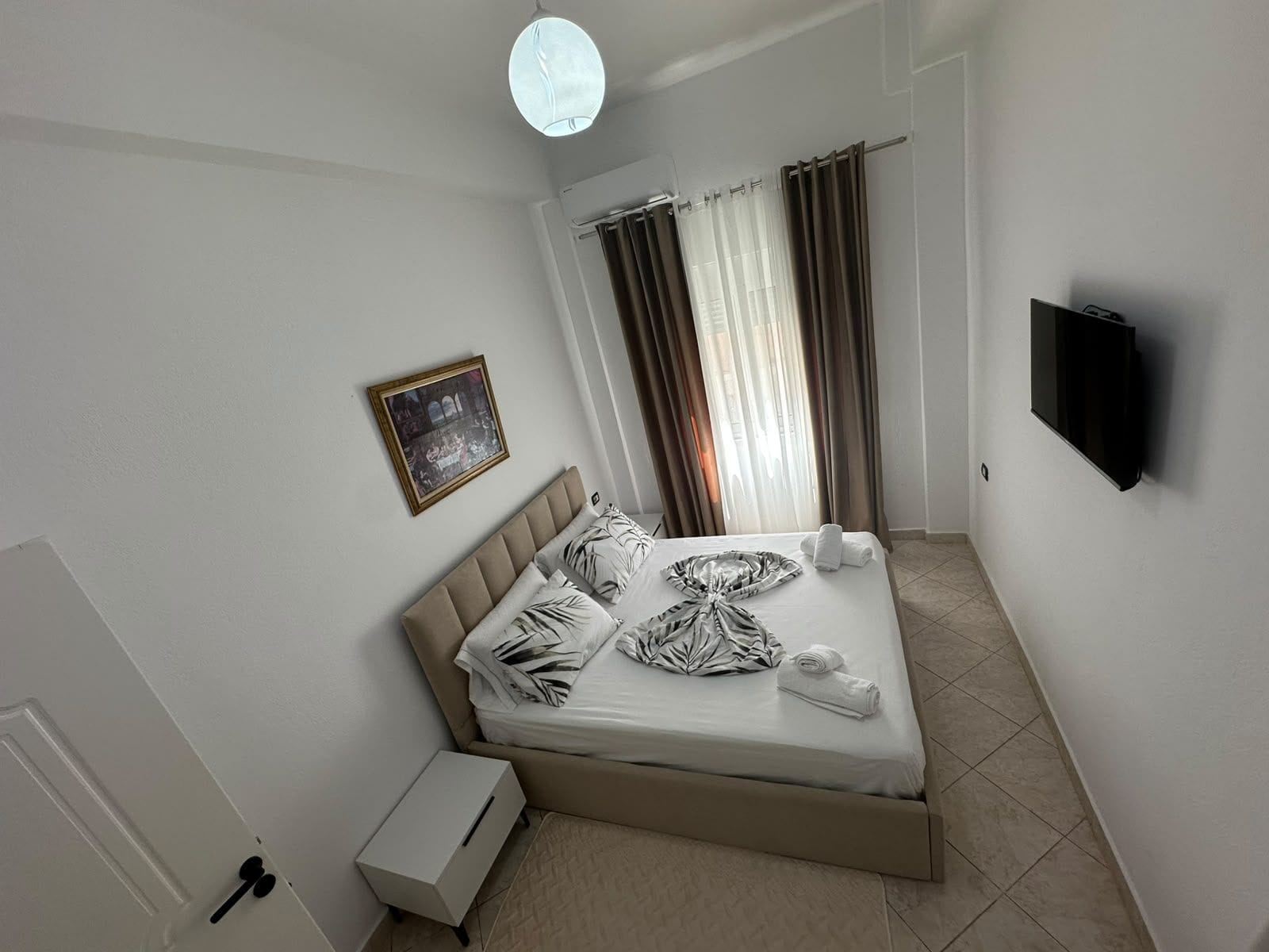 Habitación de hotel con cama, televisor de pantalla plana, un cuadro en la pared y ventana con cortinas.