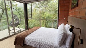 A modern bedroom with a large bed, a view of greenery through large windows, and a wooden headboard. - Samay Forest (Quito)