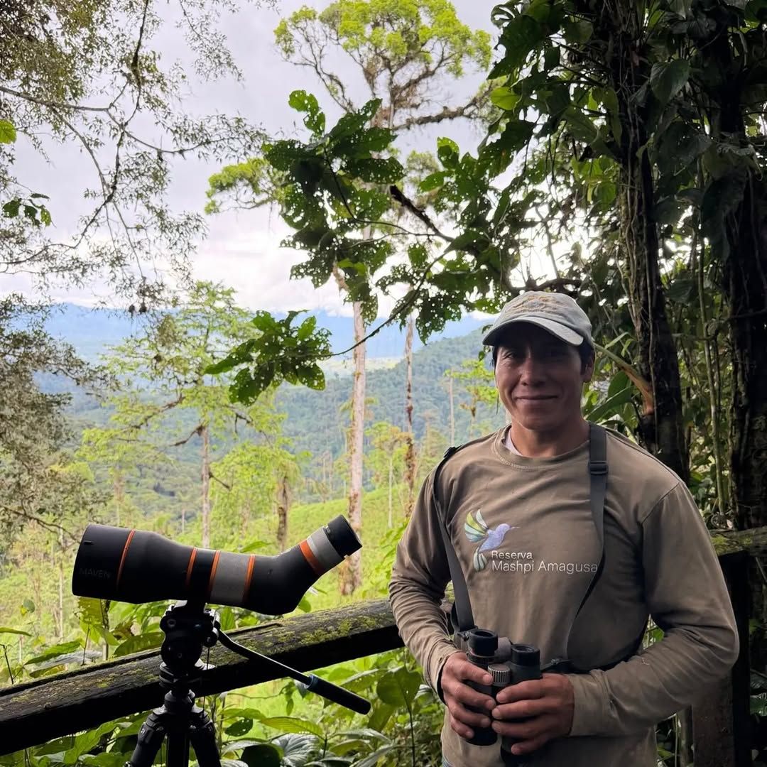 Una persona de pie junto a un telescopio, usando un gorro y una camiseta con un logotipo, en una zona boscosa con árboles y una montaña al fondo.
