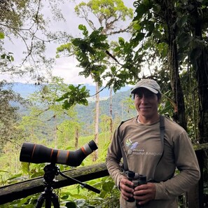 A person standing next to a telescope, wearing a cap and a shirt with a logo, in a forested area with trees and a mountain in the background. - Samay Forest (Pacto)
