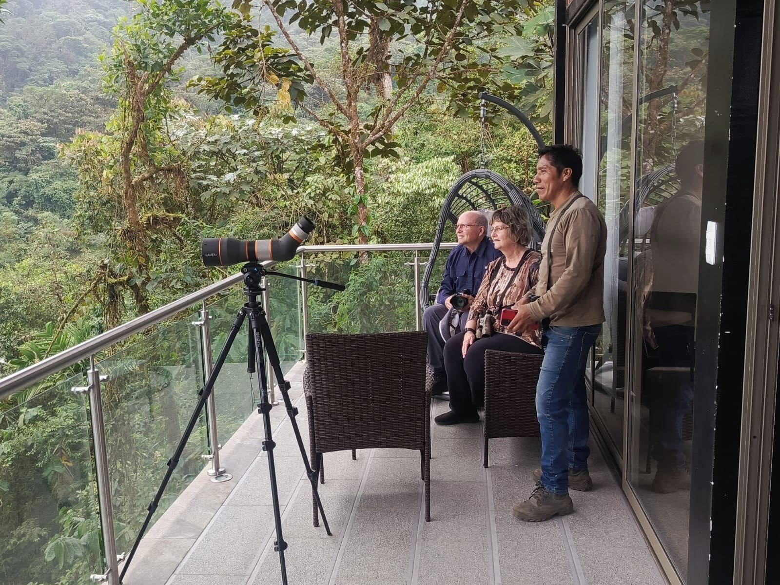 A balcony with a telescope, wicker furniture, and a glass railing overlooking a forested area.