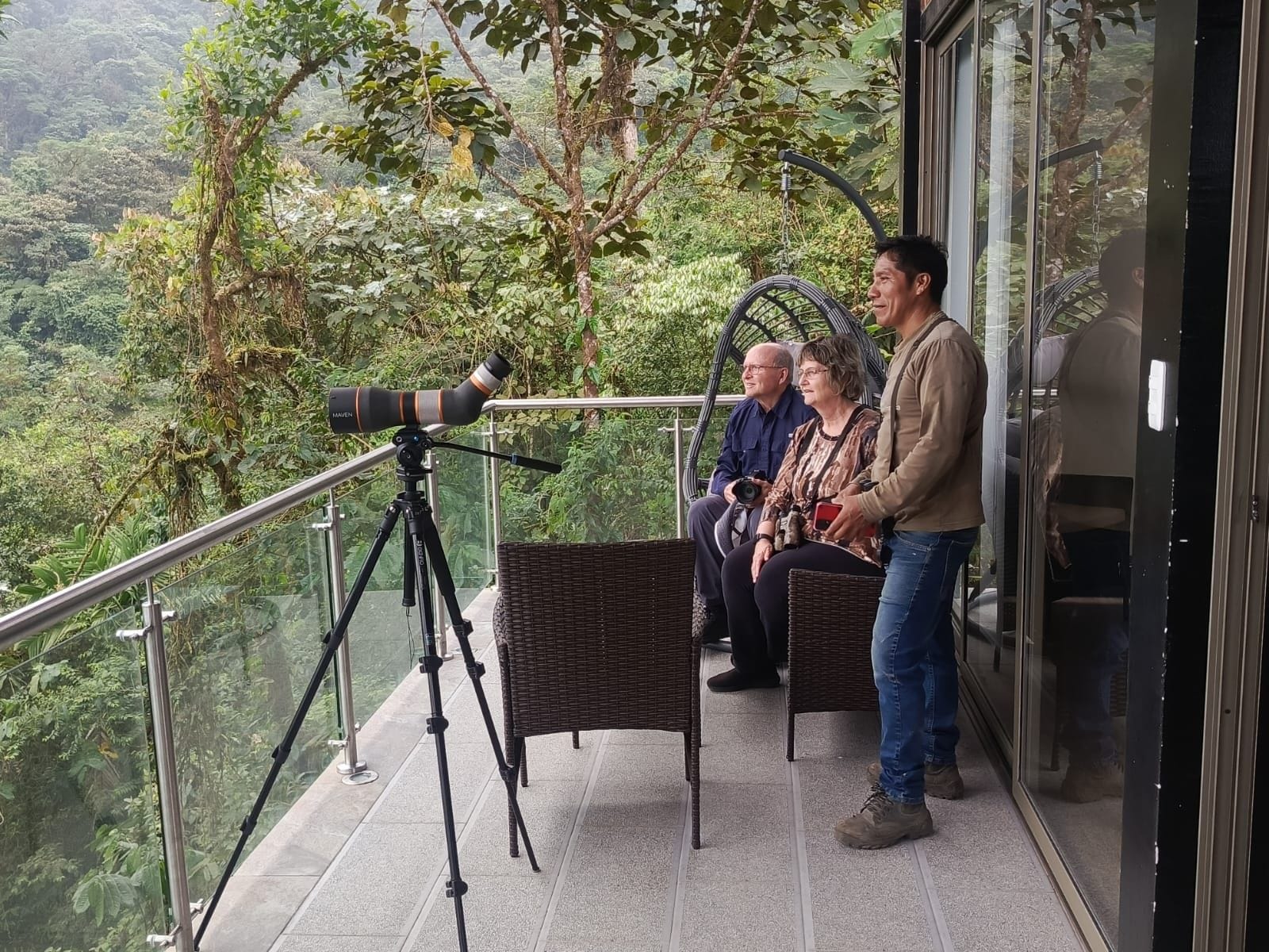A balcony with a telescope, wicker furniture, and a glass railing overlooking a forested area.