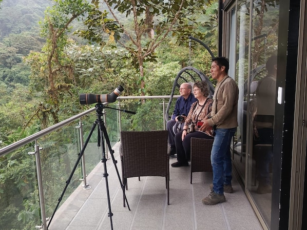Un balcón con un telescopio, muebles de mimbre y barandilla de vidrio con vista a un área boscosa.
