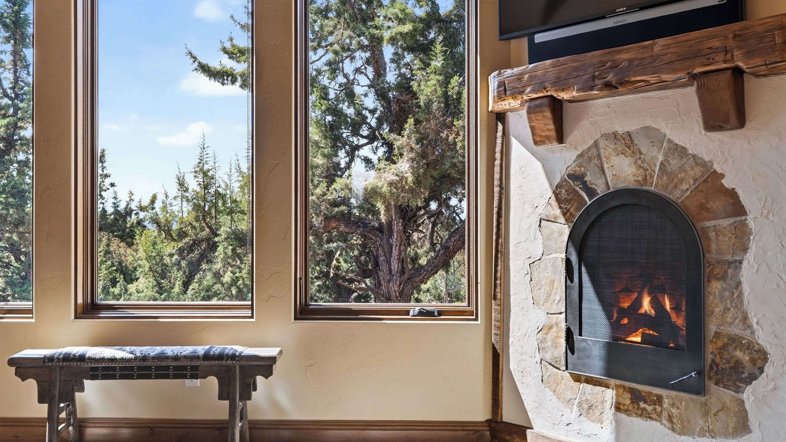 A room with a stone fireplace, large windows with a view of trees, and a bench.