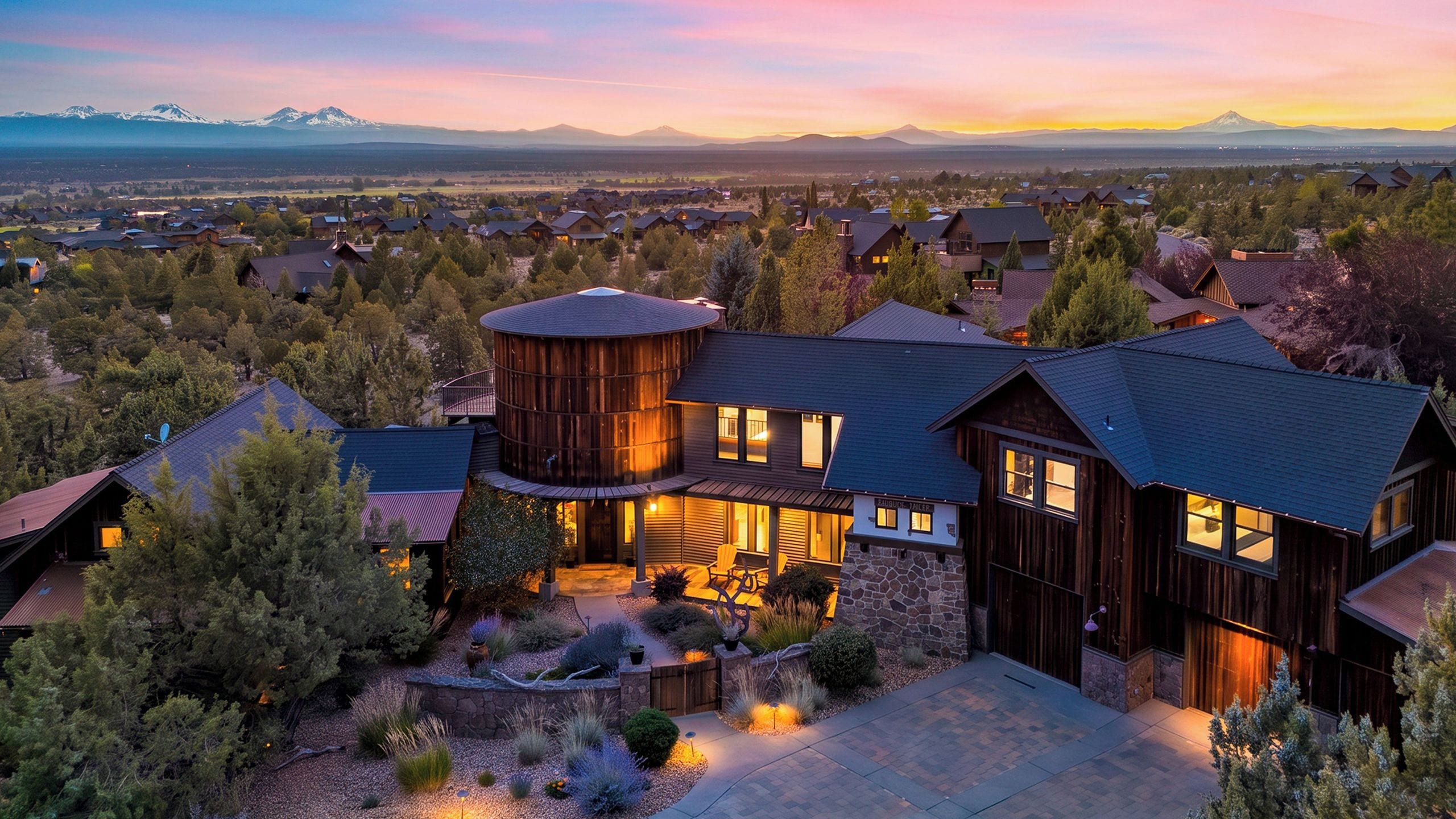 A large, multi-story house with a prominent circular tower, surrounded by a well-maintained garden and a view of distant mountains.