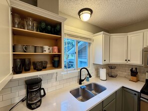 A kitchen with a built-in shelf holding various glassware, a coffee maker, and a window with a view of a house.