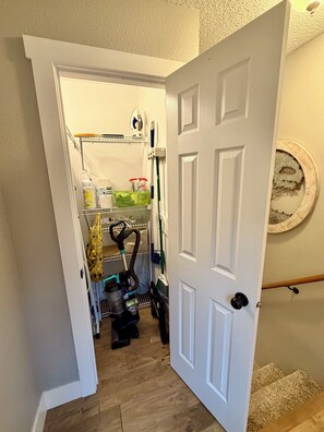 A hallway leading to a laundry room with a vacuum cleaner, ironing board, and cleaning supplies. - The River House (Bismarck)