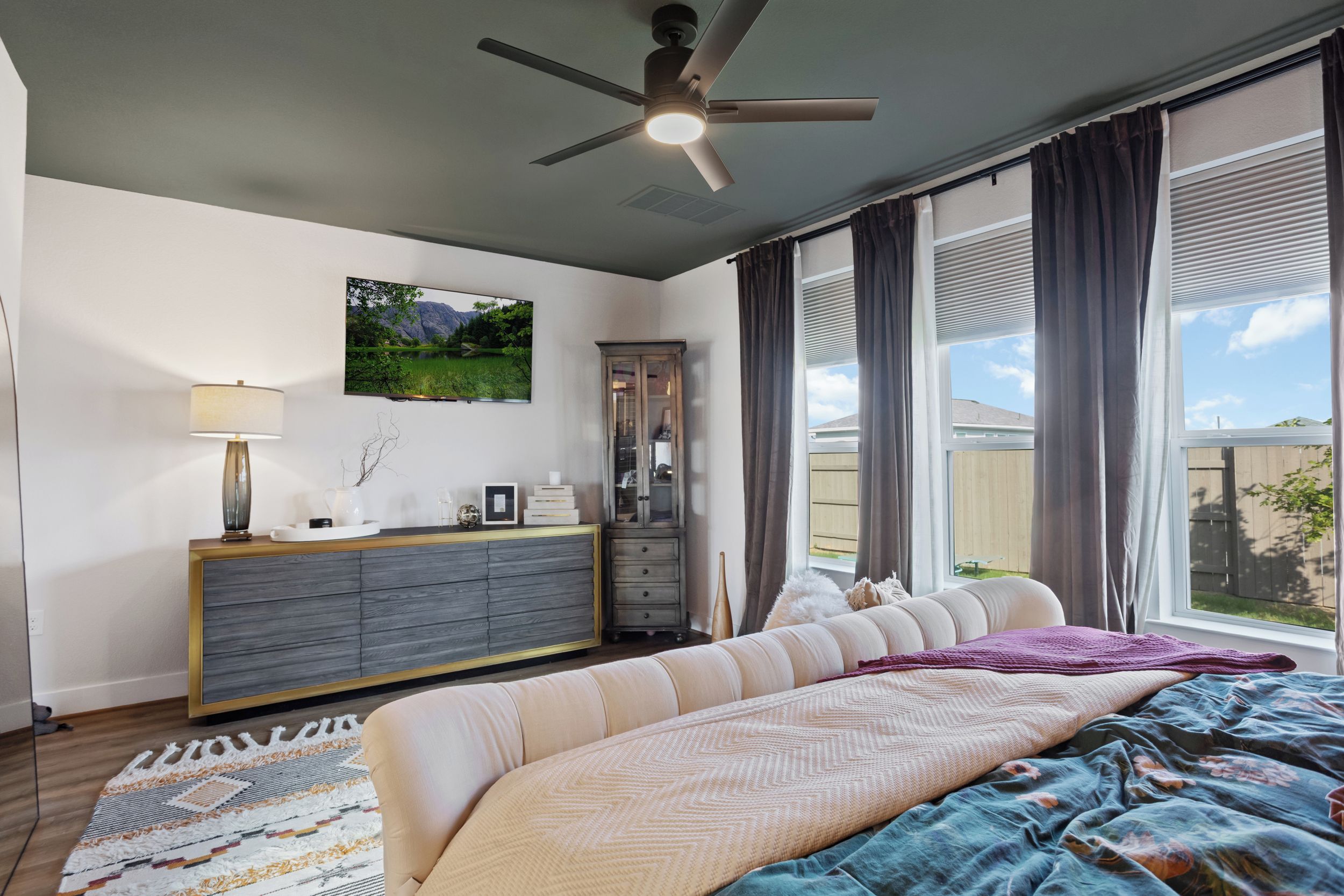 Ein Schlafzimmer mit Bett, Kommode, Deckenventilator, einem Fenster mit Vorhängen und einem Bild an der Wand.