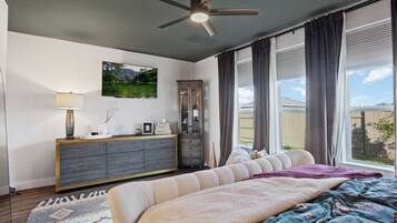 Ein Schlafzimmer mit Bett, Kommode, Deckenventilator, einem Fenster mit Vorhängen und einem Bild an der Wand.