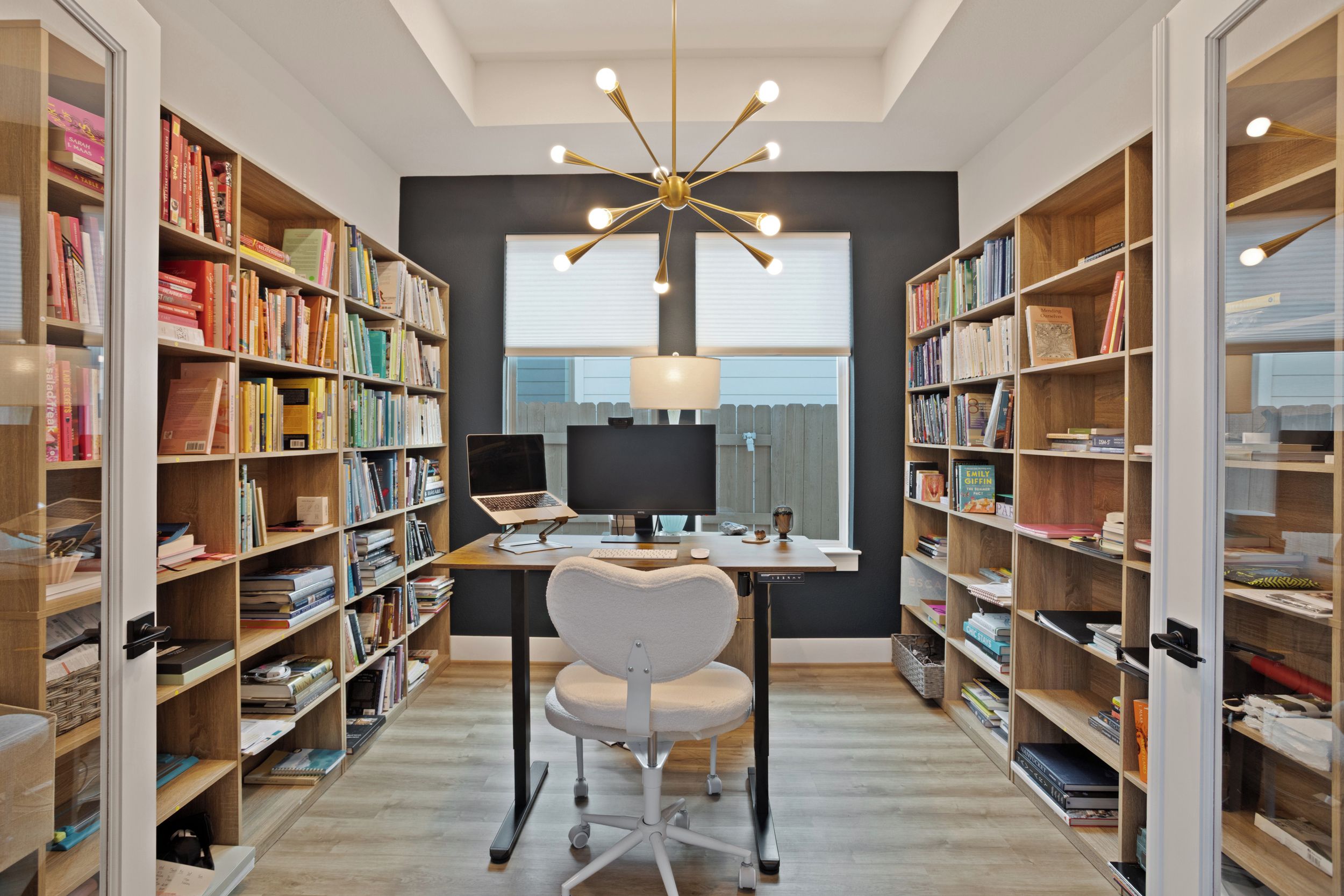 Uma biblioteca moderna com estantes de livros em ambos os lados, uma mesa com um laptop e uma luminária de teto singular.