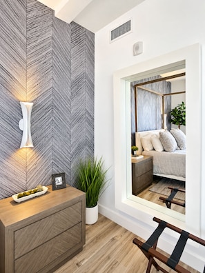 En modern inredning med en vägg som har ett fiskbensmönster, ett sängbord med en lampa, en krukväxt och en utsikt över ett sovrum genom ett stort fönster.