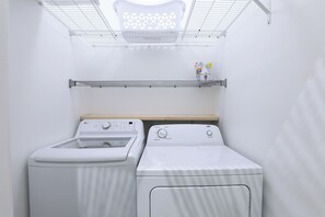 A laundry room with two white washing machines, a metal shelf with detergent bottles, and a ceiling light fixture. - The Sweet Escape Okanagan (Kelowna)