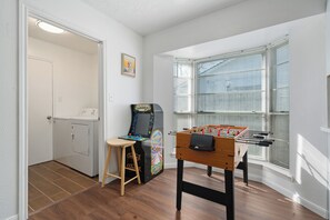 Een kamer met een wasmachine, een tafelvoetbaltafel, een raam met zonwering en een deur.
