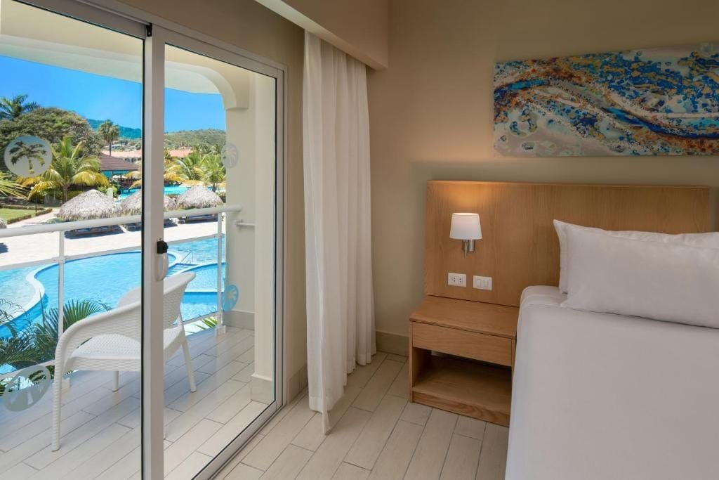 Habitación de hotel con cama, mesita de noche, lámpara, un cuadro y una puerta corrediza de vidrio que da a una zona de piscina con sillas y sombrillas.