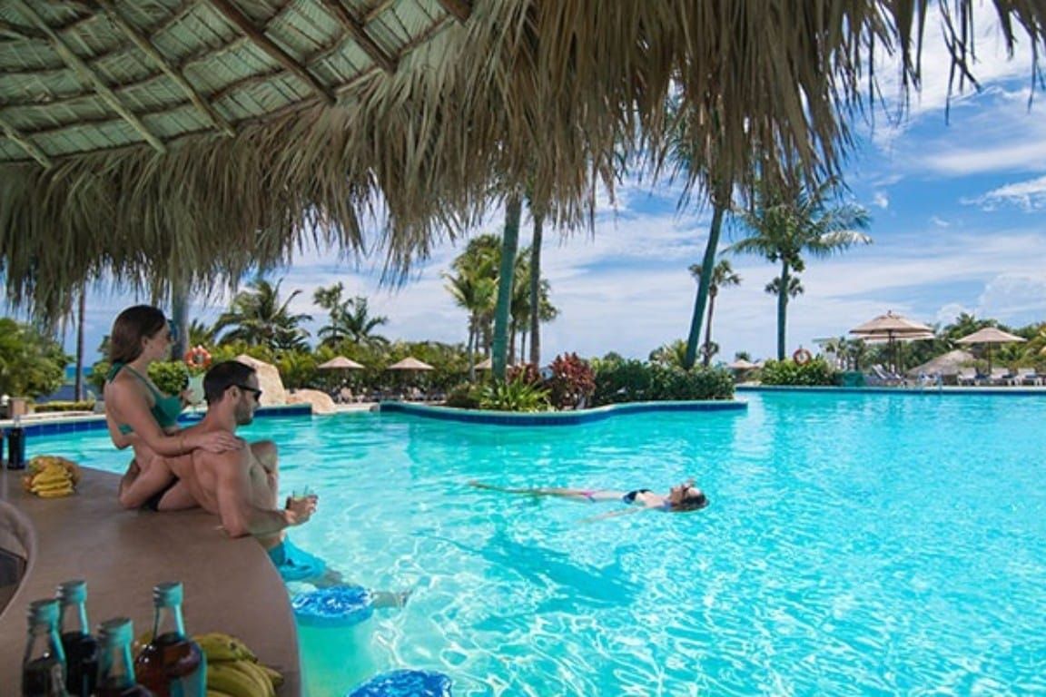A swimming pool with people relaxing and sunbathing, surrounded by palm trees and umbrellas.