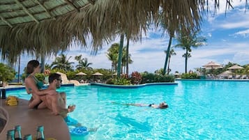Piscina com pessoas relaxando e tomando sol, cercada por palmeiras e guarda-sóis.