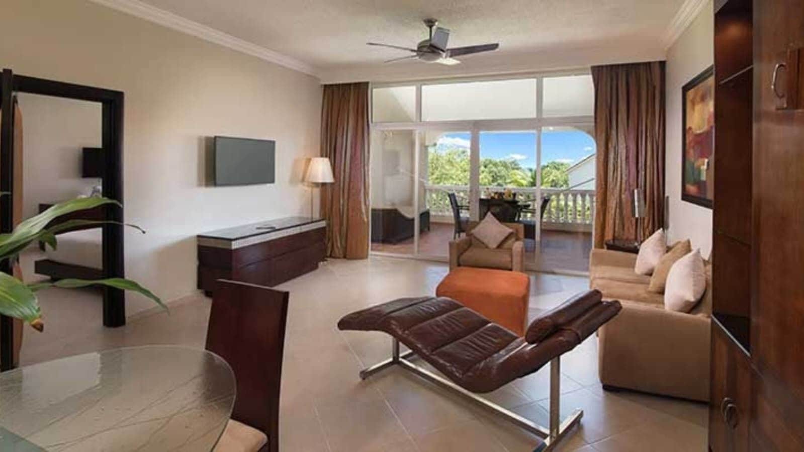 A hotel room with a balcony, a sofa, a recliner, a TV, and a desk.