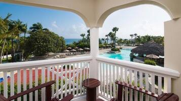 A balcony with a view of a pool and the ocean, surrounded by palm trees and lounge chairs.