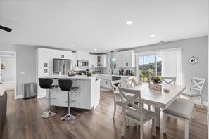 A modern kitchen with white cabinetry, a central island with bar stools, and a dining area with a view of the outdoors. - 12-Acre Nashville Retreat - Sleeps 12 - Game Room (Goodlettsville)