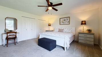 A bedroom with a bed, a desk, a chair, a ceiling fan, a bedside table with a lamp, and a wall-mounted picture.