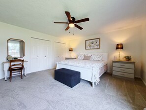 A bedroom with a bed, a desk, a chair, a ceiling fan, a bedside table with a lamp, and a wall-mounted picture.