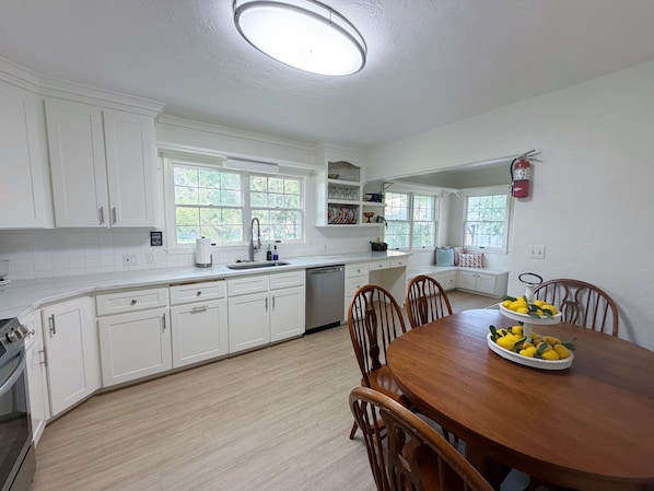 A kitchen with white cabinets, a wooden dining table with chairs, and a window with a view of greenery.