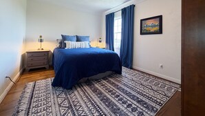 Quarto com cama, mesa de cabeceira, luminária, ventilador de teto, janela com cortinas e um quadro na parede.