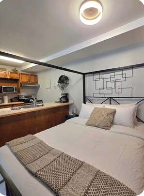 A modern bedroom with a bed, a bedside table, a wall-mounted artwork, and a kitchenette area with a microwave and coffee maker. - Panorama Mountain Escape Ski In/Out/Near Hot Tubs (Panorama)