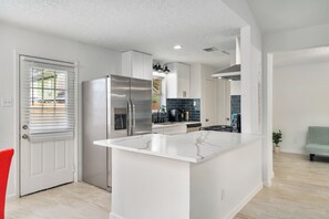 A modern kitchen with a central island, stainless steel appliances, and a view into the adjacent living area. - Asiantown Escape - Cozy 3br Bellaire Home W Game Room (Houston)