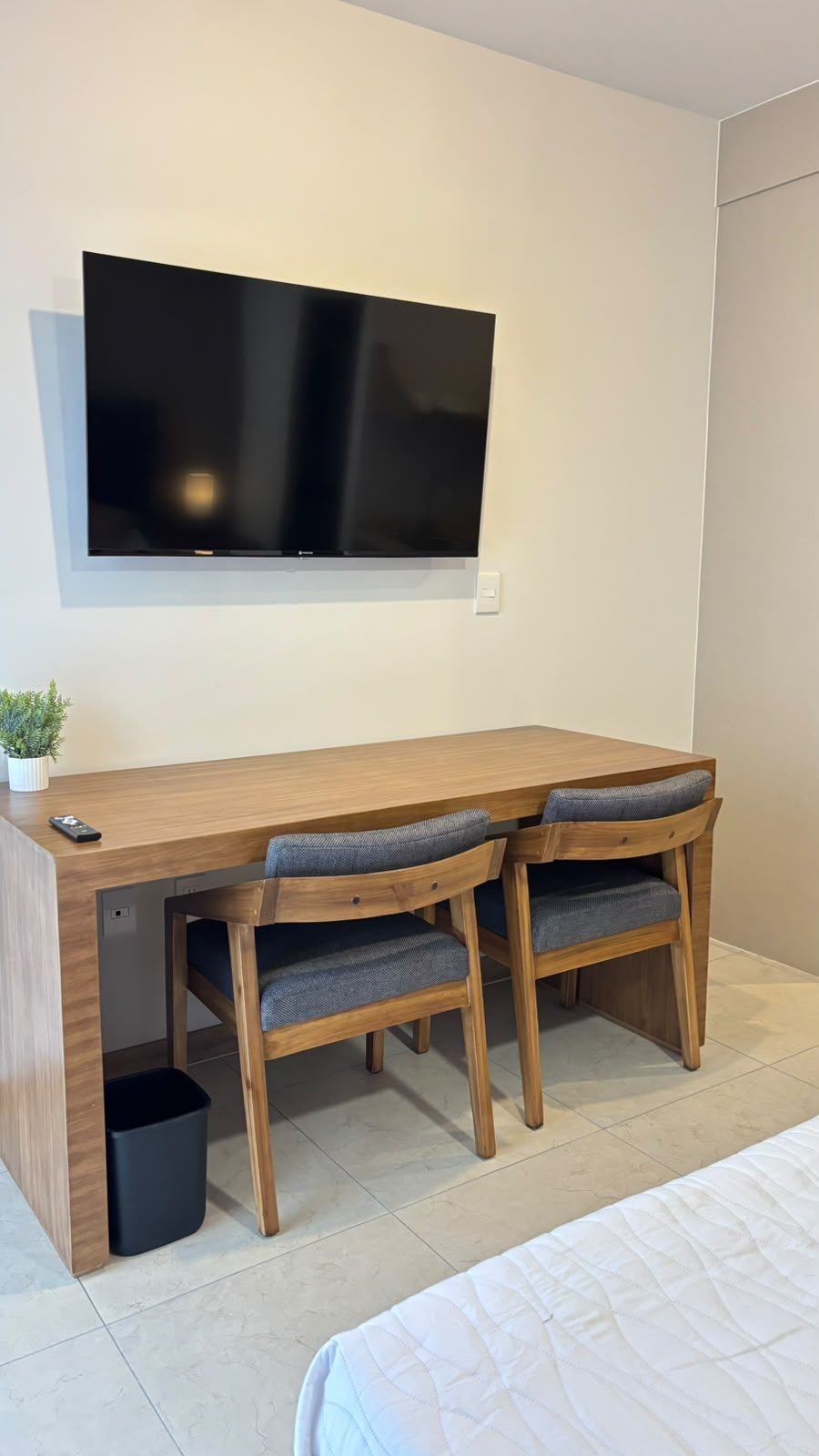 Un dormitorio con cama, un escritorio con dos sillas, un televisor montado en la pared y una pequeña planta en maceta.