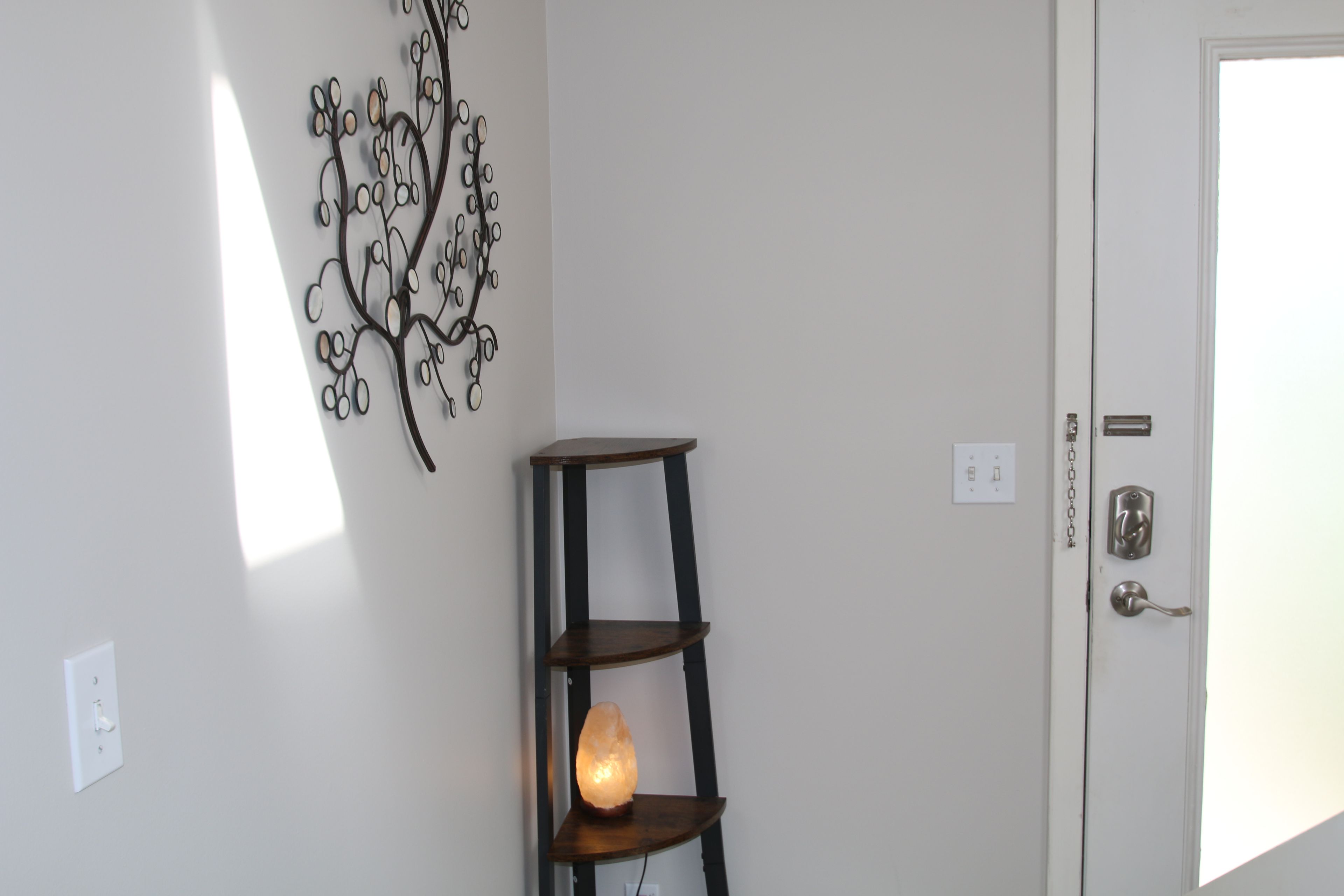 A room with a white door, a wall-mounted tree decoration, and a wooden shelf with a light.