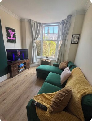 A living room with a television, a green sofa, a yellow blanket, and a window with curtains. - Stylish 3 Bedroom Chelmsford Sleeps 6 (Chelmsford)