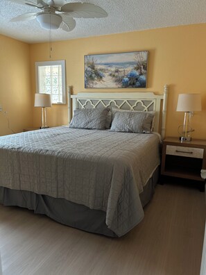A bedroom with a bed, nightstands, a ceiling fan, a window with a view, and a painting on the wall.