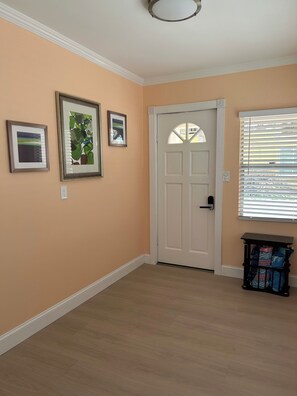 A room with peach walls, a white door, and a window with blinds. There are three framed pictures on the wall. - Beautiful 2-bedroom Cottage across the street from the beach. (Indian Shores)