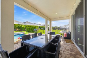 A covered patio with a glass table and wicker chairs, a barbecue grill, and a swimming pool in the background. - 10BR near Disney, sleeps 28, pool, spa & theater (Kissimmee)