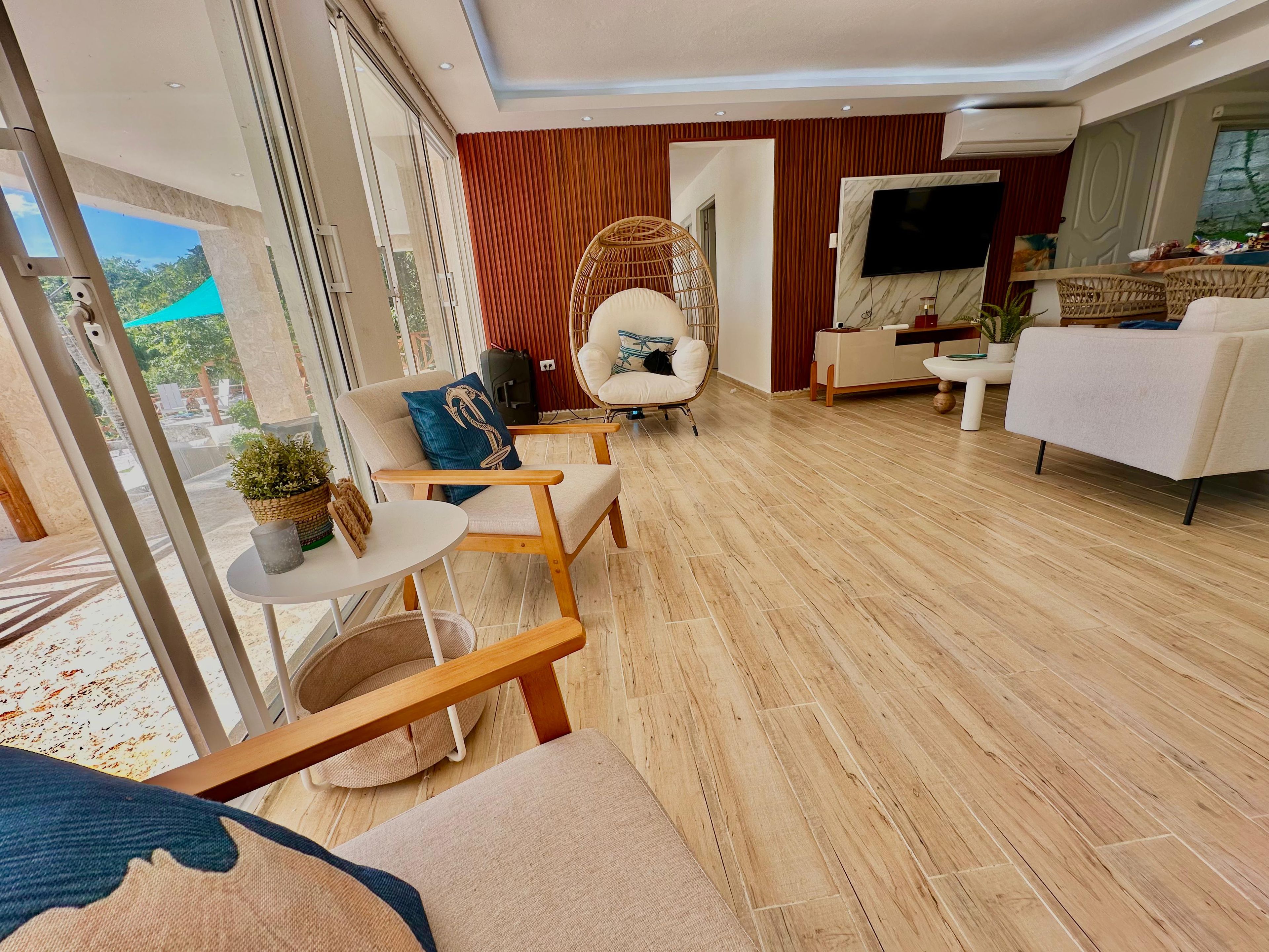 Ein modernes Wohnzimmer mit einer Couch, Sesseln, einem Fernseher und Blick durch große Fenster.
