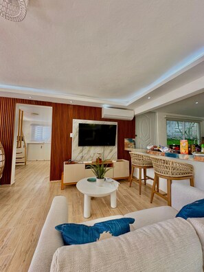 A modern living room with a flat-screen TV, a dining area with wicker chairs, and a kitchen visible through an open door.