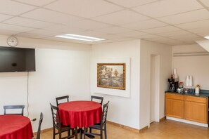 Ein Zimmer mit zwei runden Tischen, die mit roten Tischdecken bedeckt sind, einer Uhr an der Wand, einem gerahmten Gemälde und einer Küchenzeile mit einer Kaffeemaschine und Tassen.