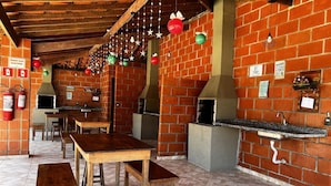 A brick-walled outdoor dining area with wooden tables and benches, a brick oven, and hanging decorations. - Ap p/ família, 3 quartos, 5min da praia (Ubatuba)