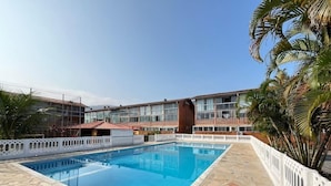 A swimming pool with a stone deck, surrounded by a white fence, and a building with multiple windows in the background. - Ap p/ família, 3 quartos, 5min da praia (Ubatuba)