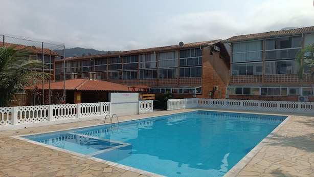 A swimming pool with a stone deck, a white fence, and a building with a tiled roof in the background. - Ap p/ família, 3 quartos, 5min da praia (Ubatuba)
