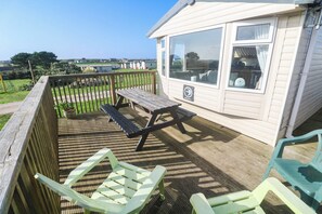A wooden deck with a picnic table and chairs, a small building with windows, and a view of a green landscape. - Sennen Sunset (Penzance)